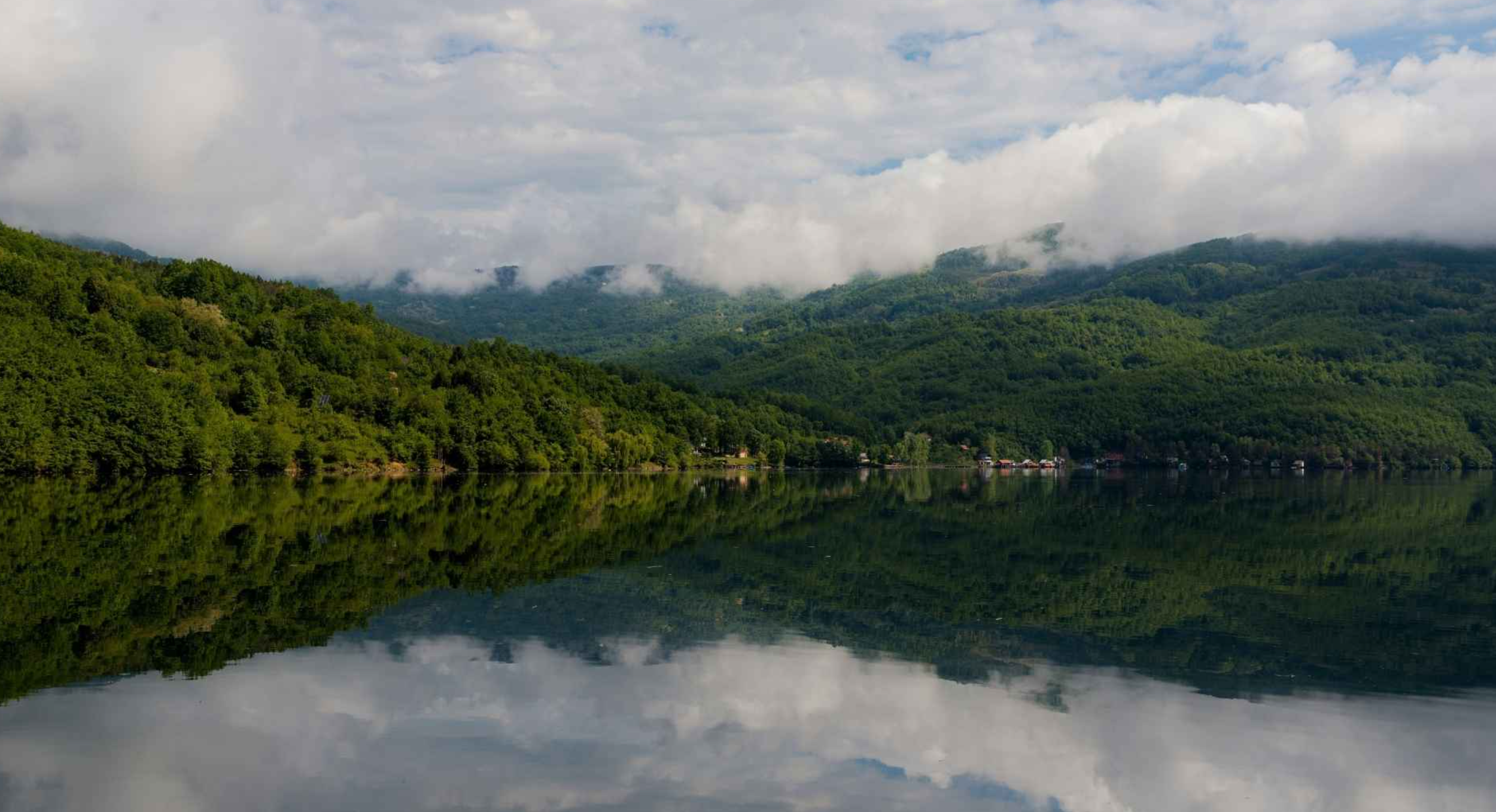 A calm lake reflecting green forested hills and partly cloudy sky.