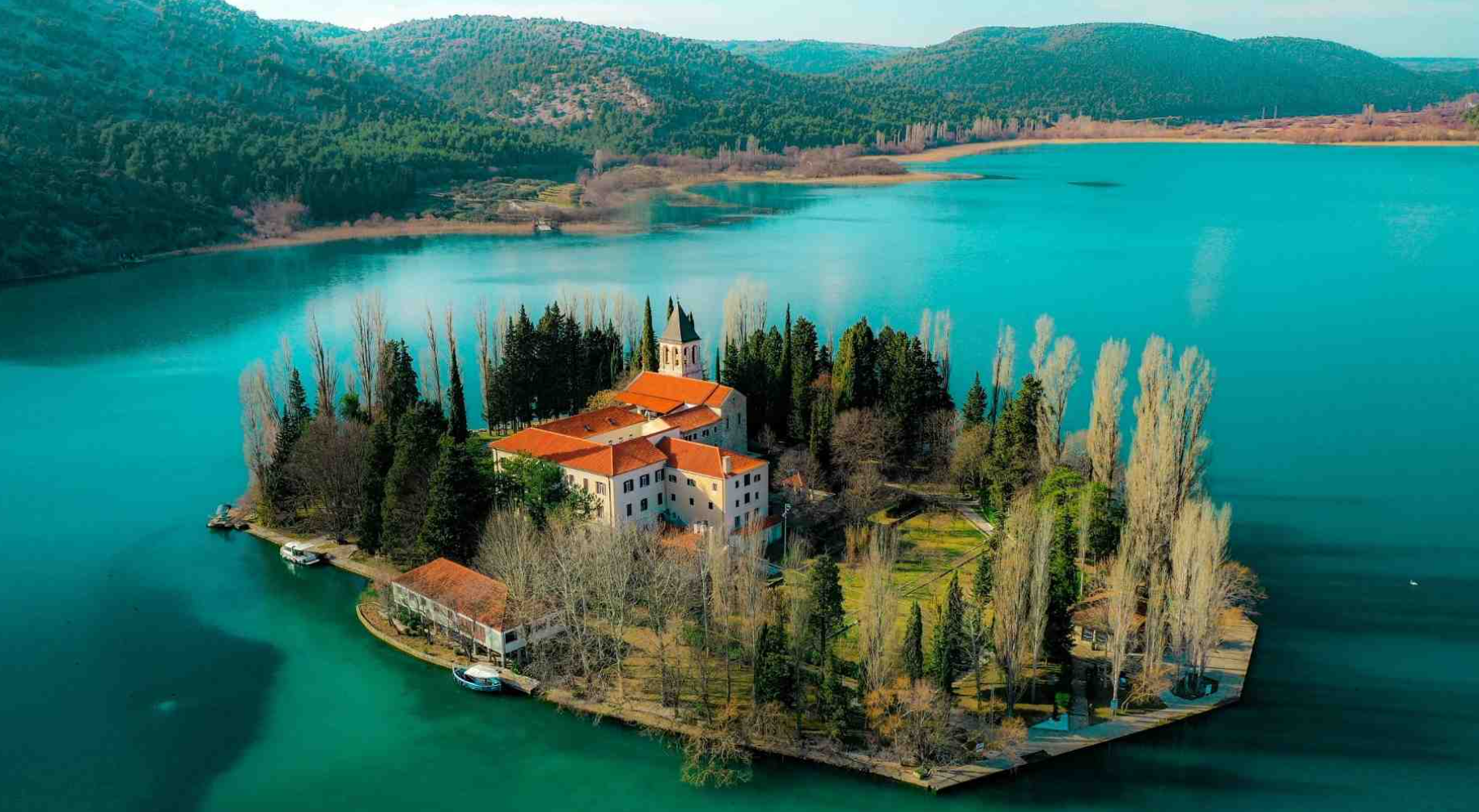 Aerial view of a small, historic church and buildings with red roofs on a narrow island surrounded by a large blue lake, with mountains in the background.