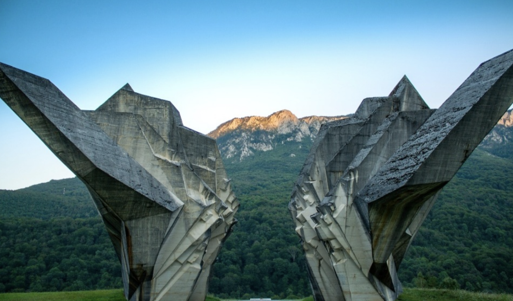 Large concrete sculpture resembling two hands clasped together, with a green forest and mountains in the background.