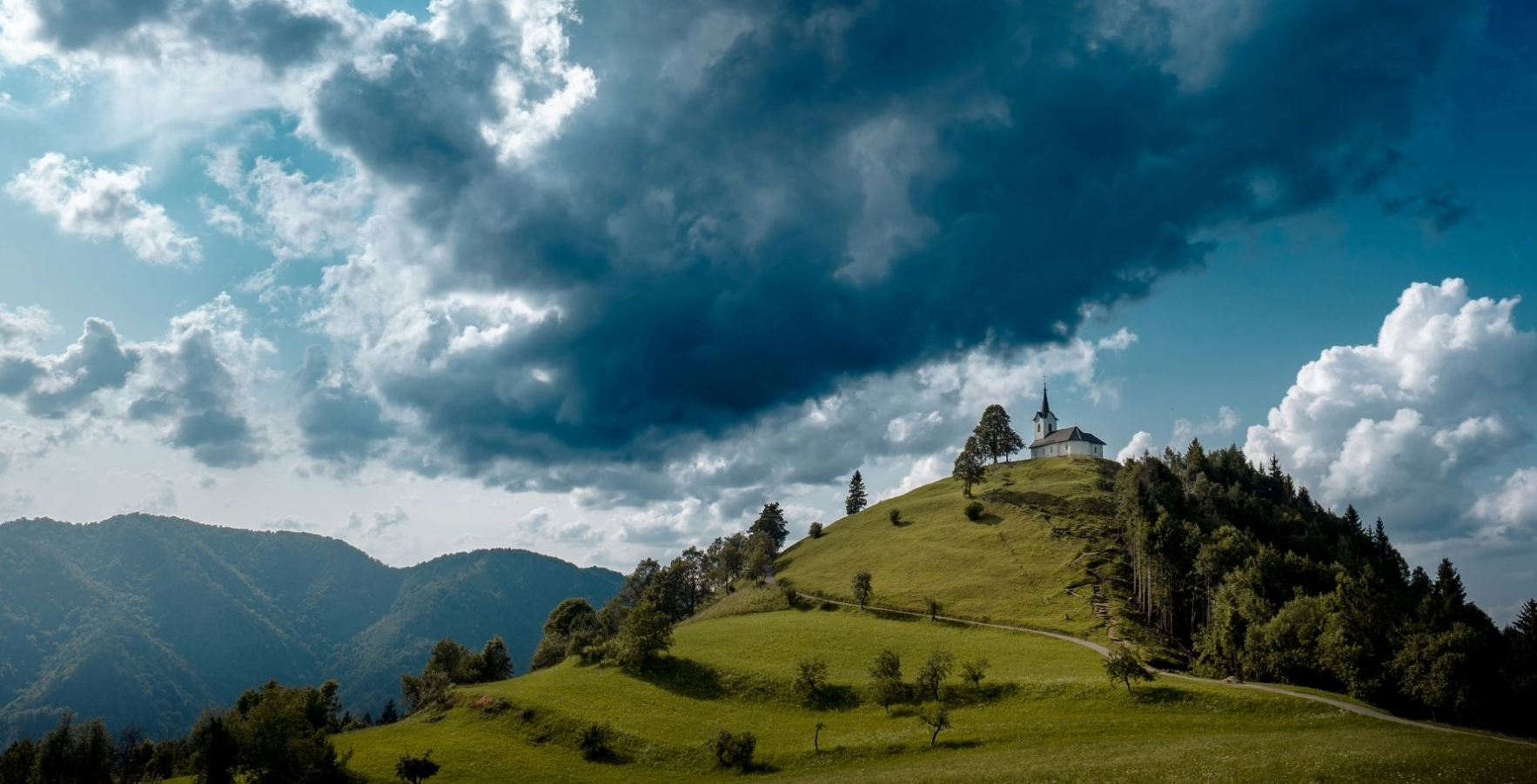 A church with a steeple on a hilltop overlooking green rolling hills and mountains, with a partly cloudy sky overhead.