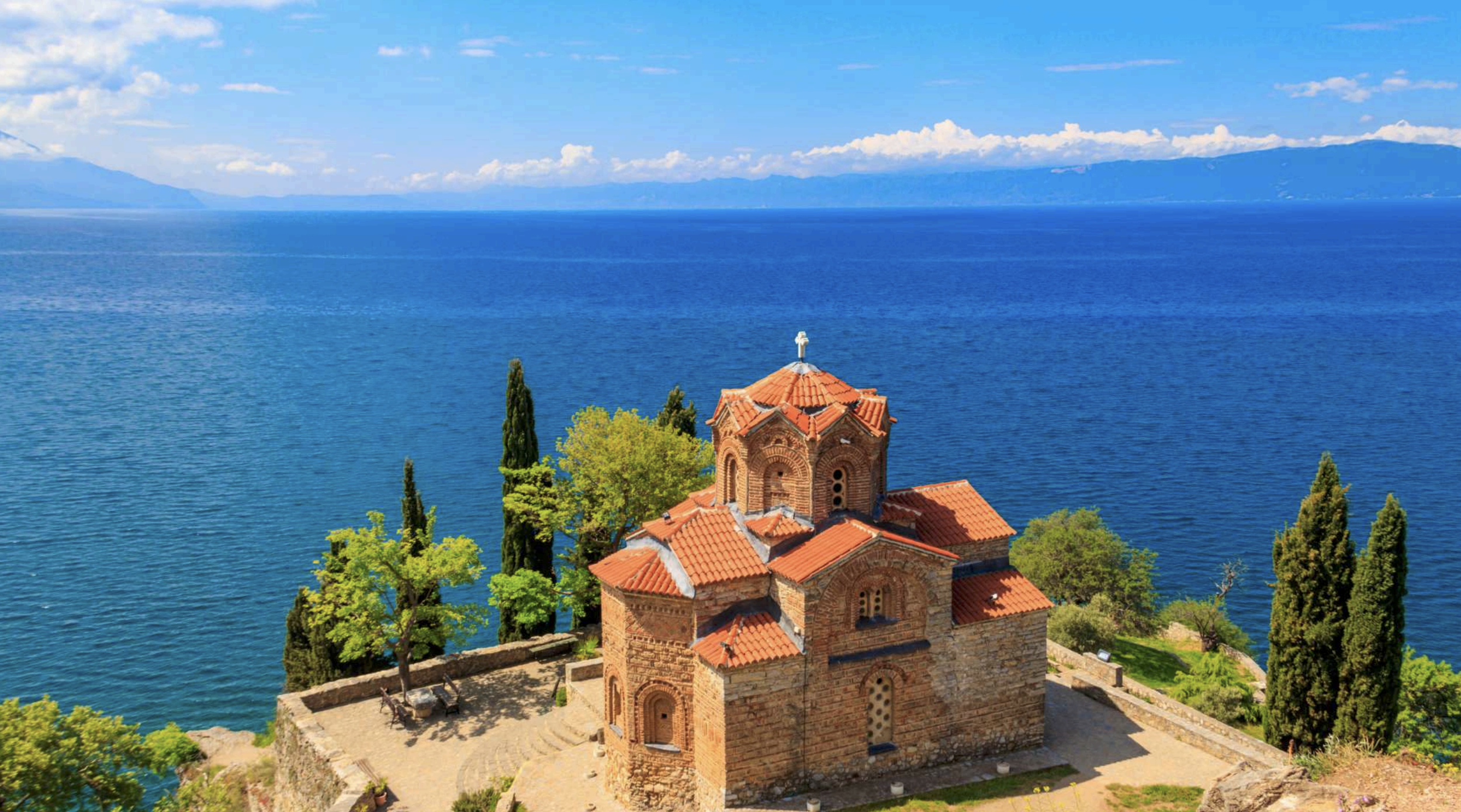A small brick church with red tile roof near a large blue lake, surrounded by green trees, with mountains in the distance, under a partly cloudy sky.