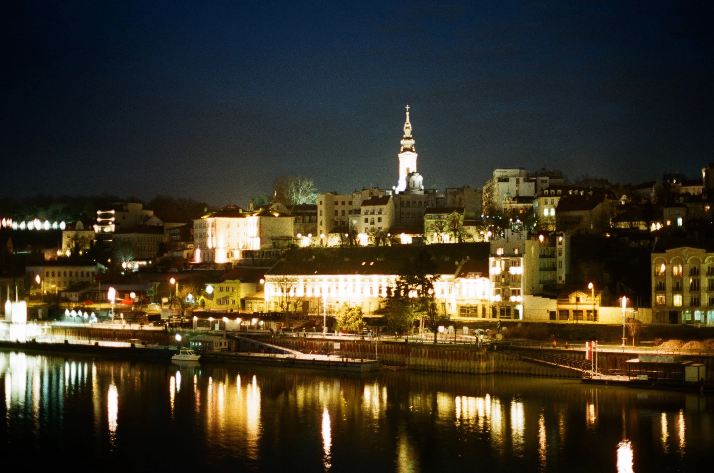 Night cityscape with illuminated buildings and church steeple across a river reflection.