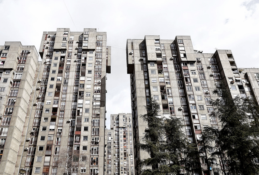 High-rise apartment buildings with many windows, balconies, and air conditioning units, under a cloudy sky, with trees in the foreground.