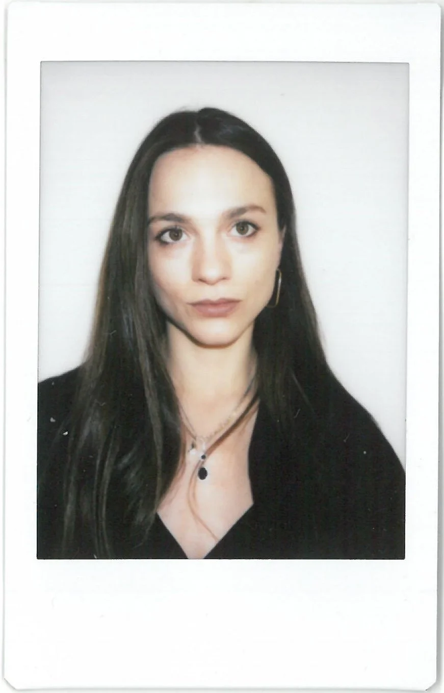 A woman with long dark hair, wearing a black top and layered necklaces, taking a portrait against a plain white background.