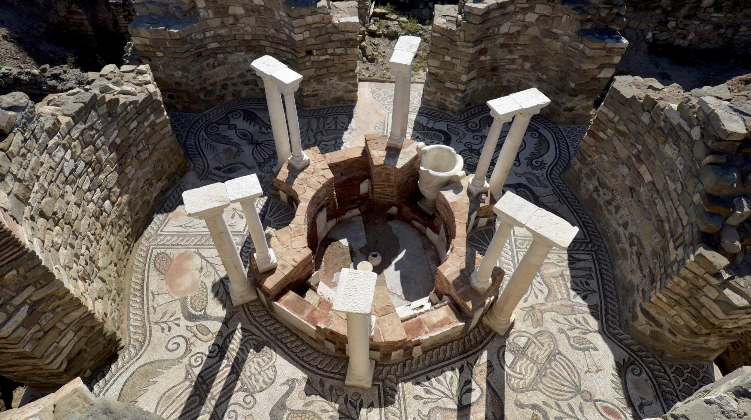 Ancient archaeological site with brick and stone structures, mosaic floor, and white columns surrounding a small round brick structure with a white marble vessel on top.