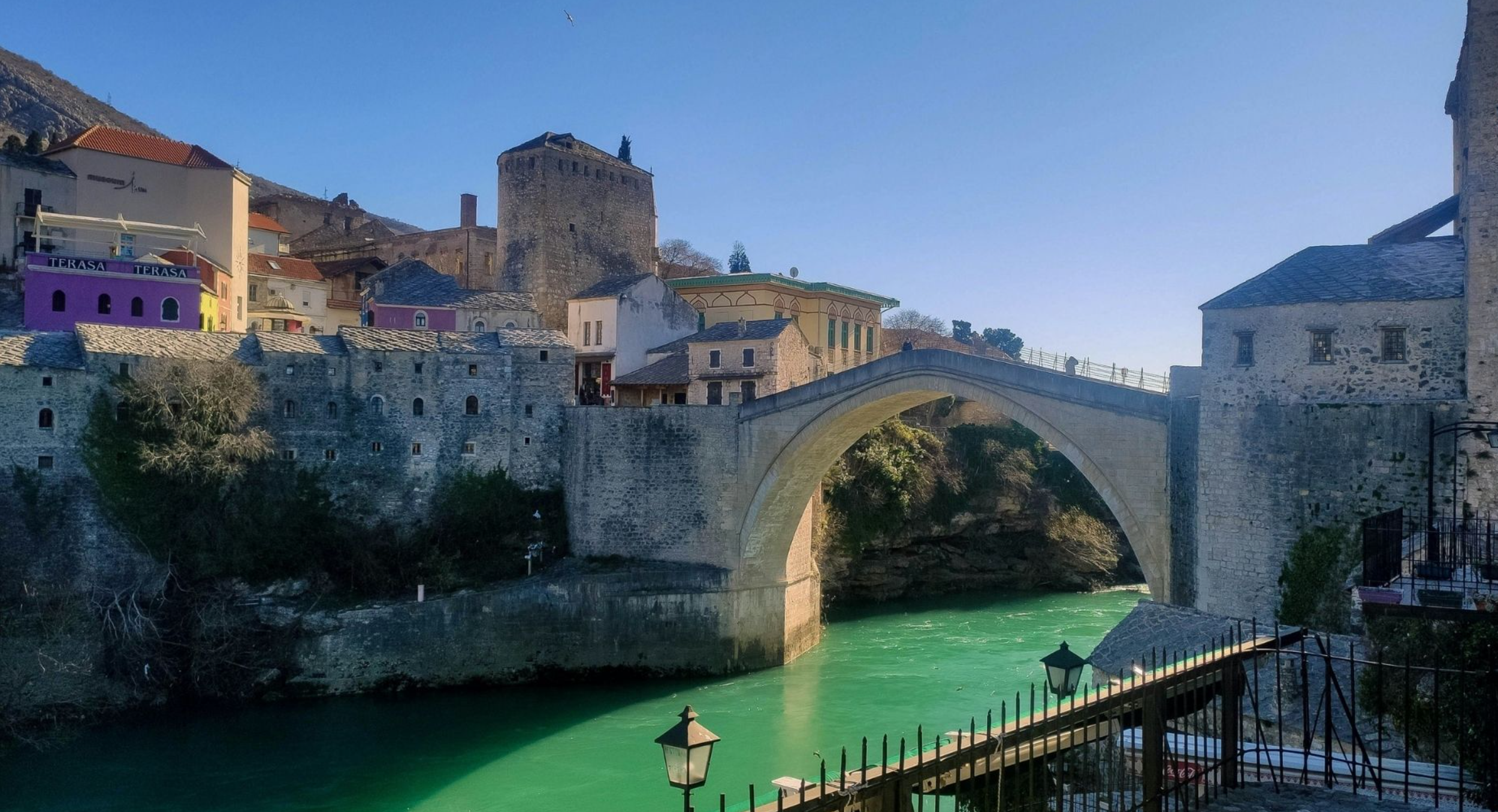 A historic stone bridge over a river, with colorful old buildings and a castle on a hill in the background, under a clear blue sky.