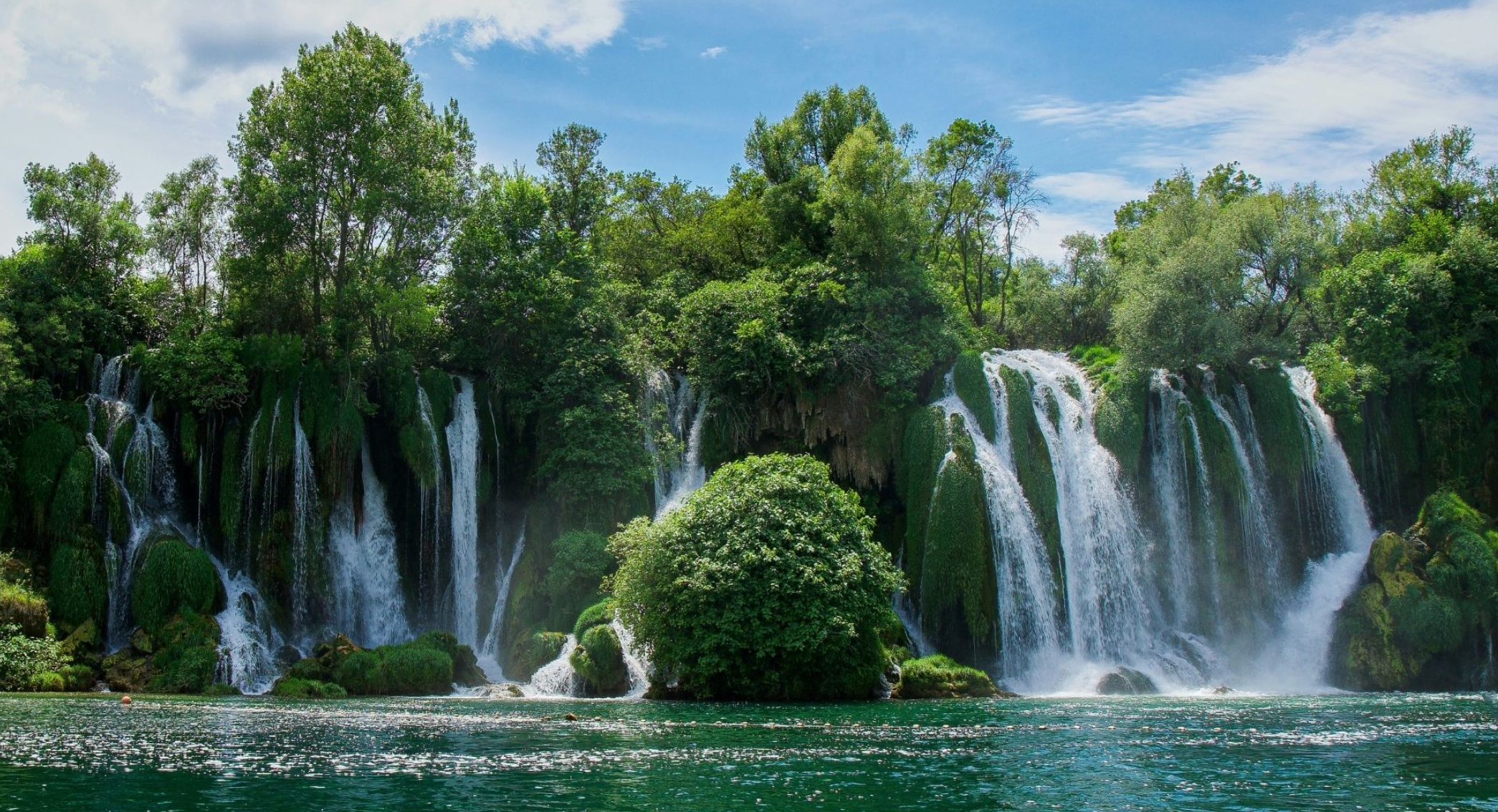 A scenic view of a lush green forest with multiple waterfalls cascading down moss-covered rocks into a calm lake below, under a partly cloudy sky.