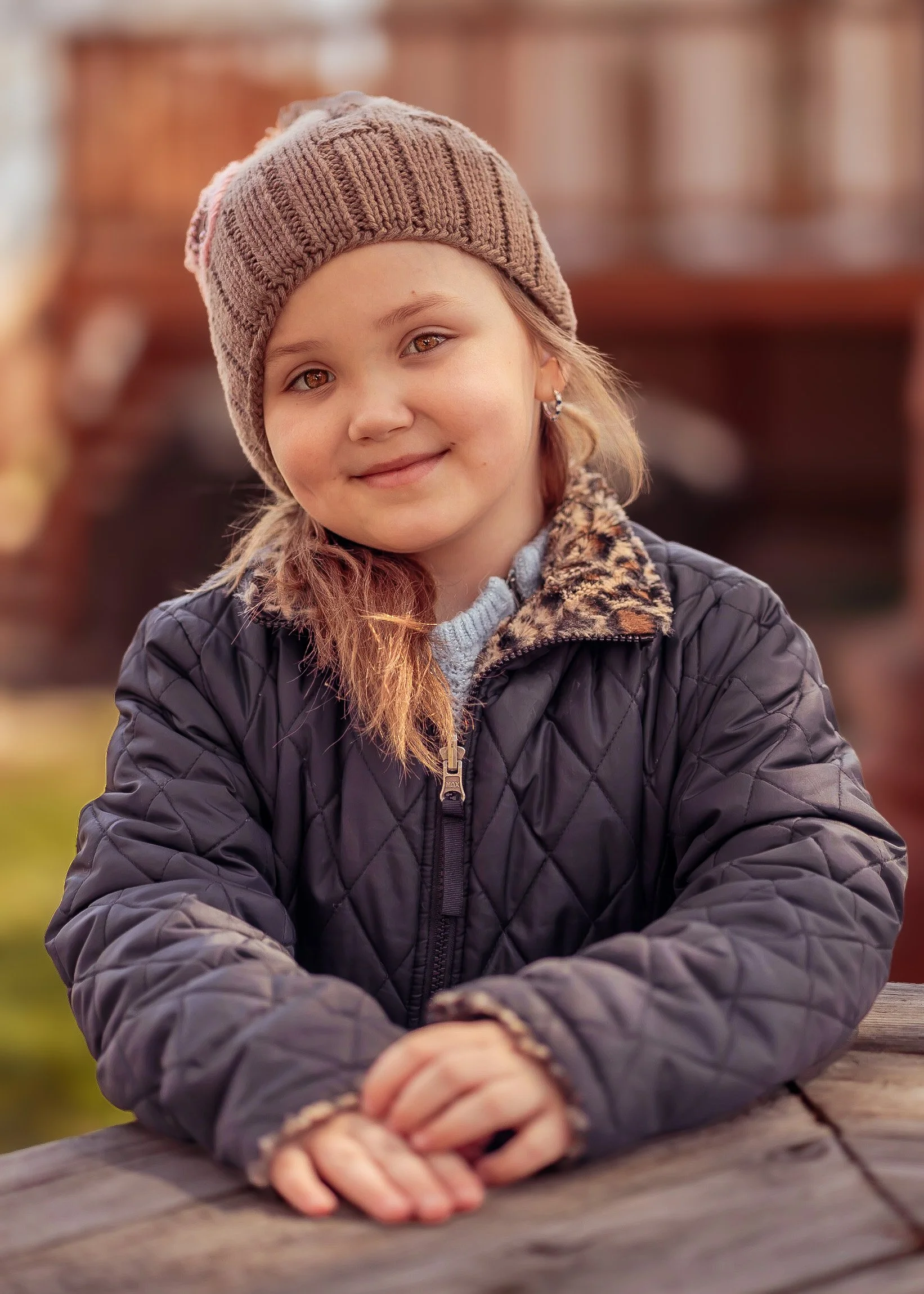 A young girl with a gentle smile, wearing a brown knit beanie, a black quilted jacket with a leopard print collar, and a gray sweater underneath, sitting outdoors at a wooden table.