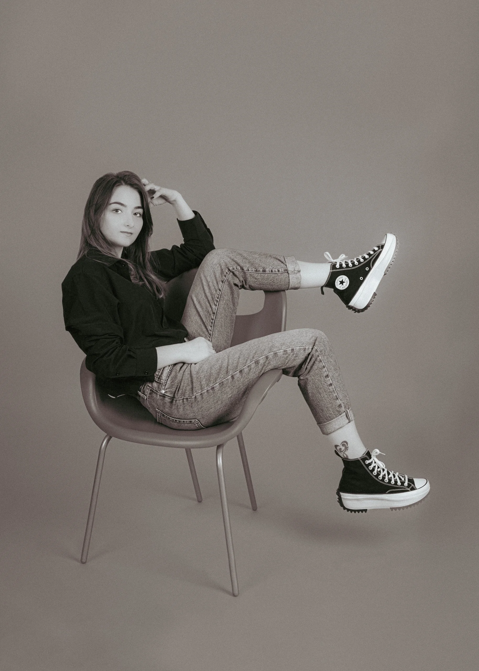 A woman sitting on a modern chair with her legs extended, wearing casual clothes including a black jacket, jeans, and black sneakers with white soles, in a minimalistic setting.