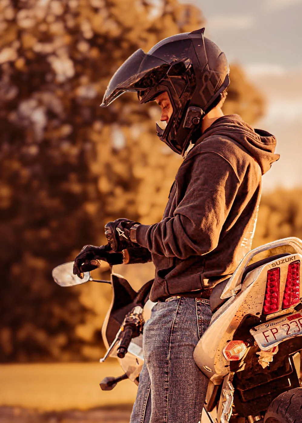 Young man wearing a motorcycle helmet and gloves, standing next to a Suzuki motorcycle at sunset.