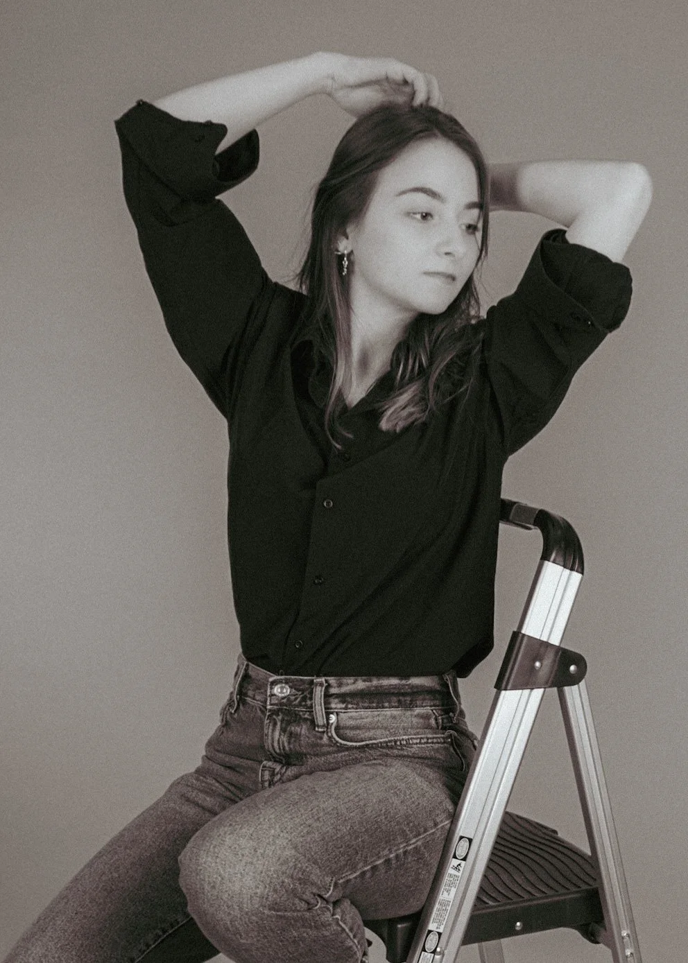Black and white photo of a woman with long hair wearing a black shirt, sitting on a small ladder with arms raised and hands behind her head.