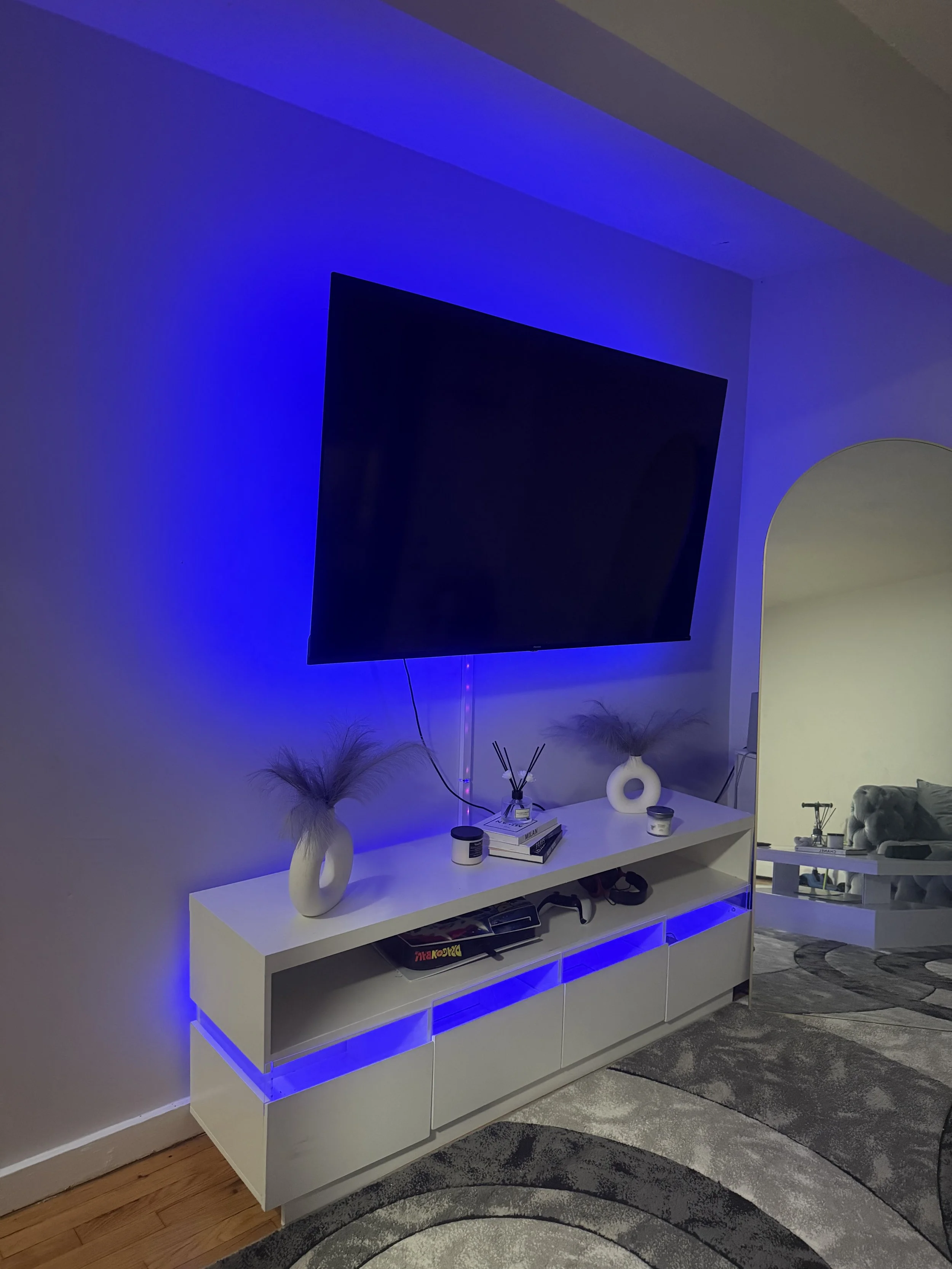 Living room with a wall-mounted flat-screen TV illuminated in blue, white TV stand with decorative vases and books, and a gray patterned rug.