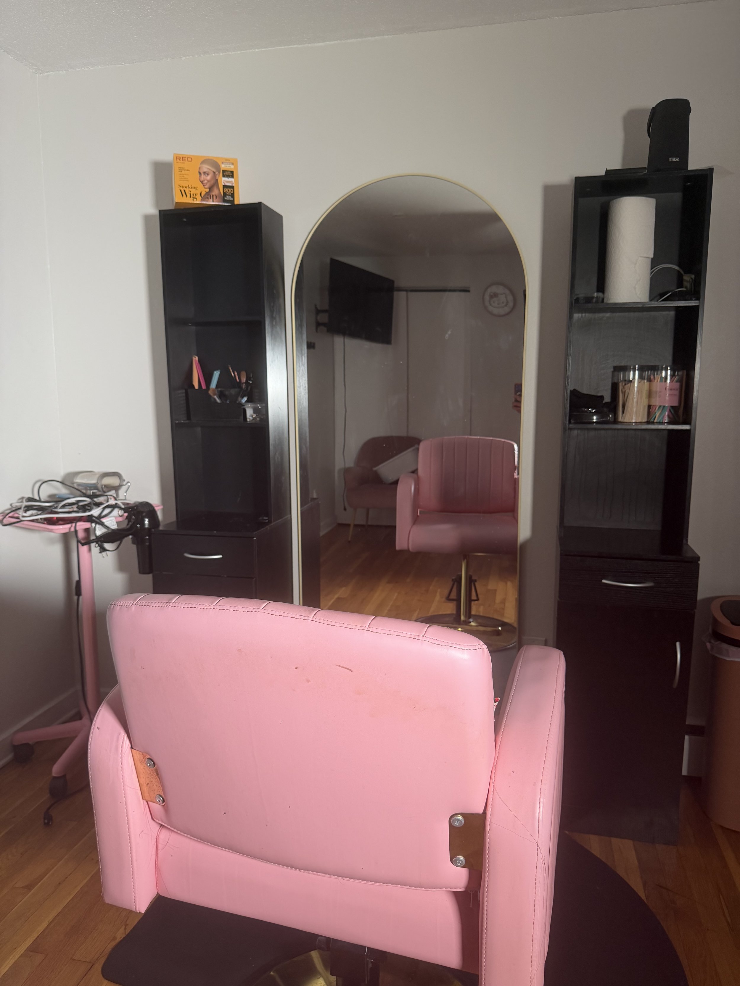 A room with a pink salon chair in the foreground, a large partially visible mirror, black shelving units with various salon tools and products, a small pink cart with tools, and a wall clock in the background.