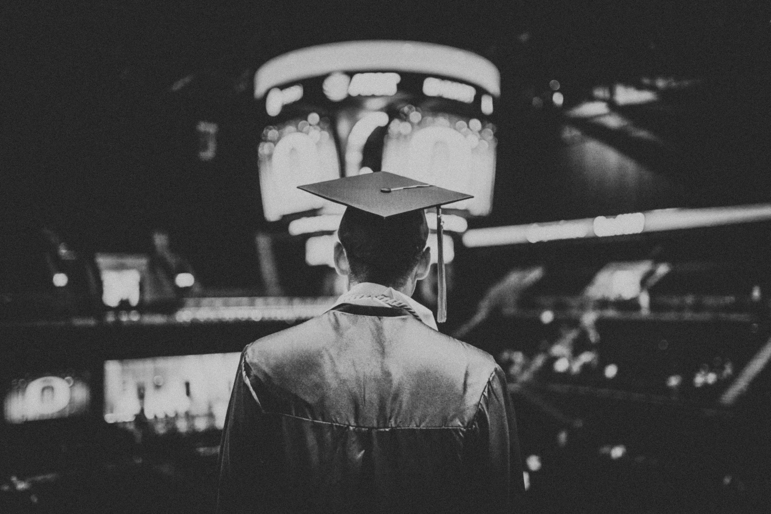 A person in a graduation cap and gown facing away, standing in front of a large, illuminated sign at night.