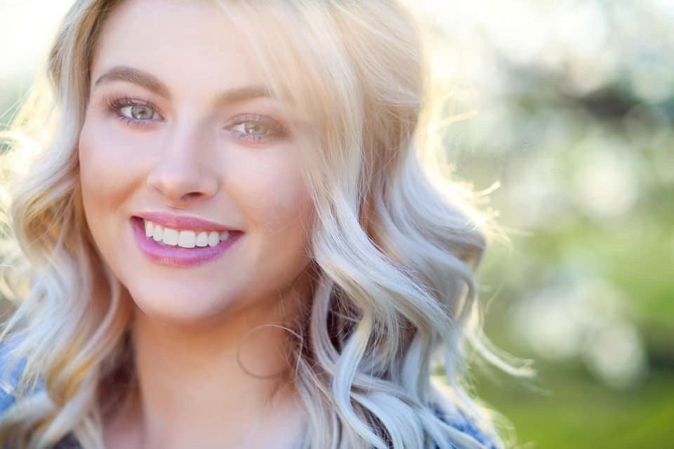 Close-up of a smiling young woman with blonde, wavy hair and blue eyes outdoors with sunlight in the background.