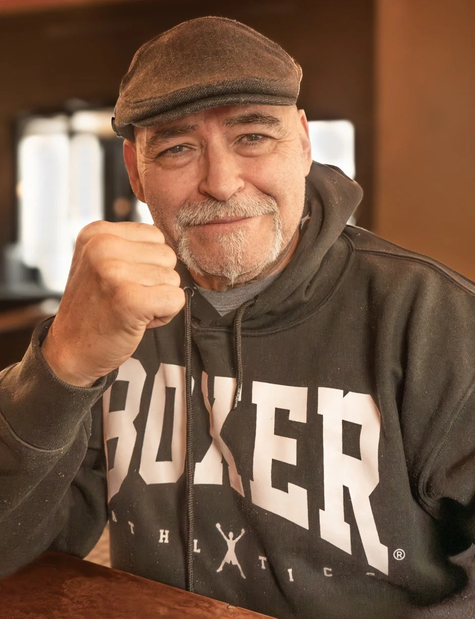 A middle-aged man with a white beard and mustache, wearing a brown cap and a black hoodie, making a fist gesture near his face and smiling inside a cozy indoor setting.