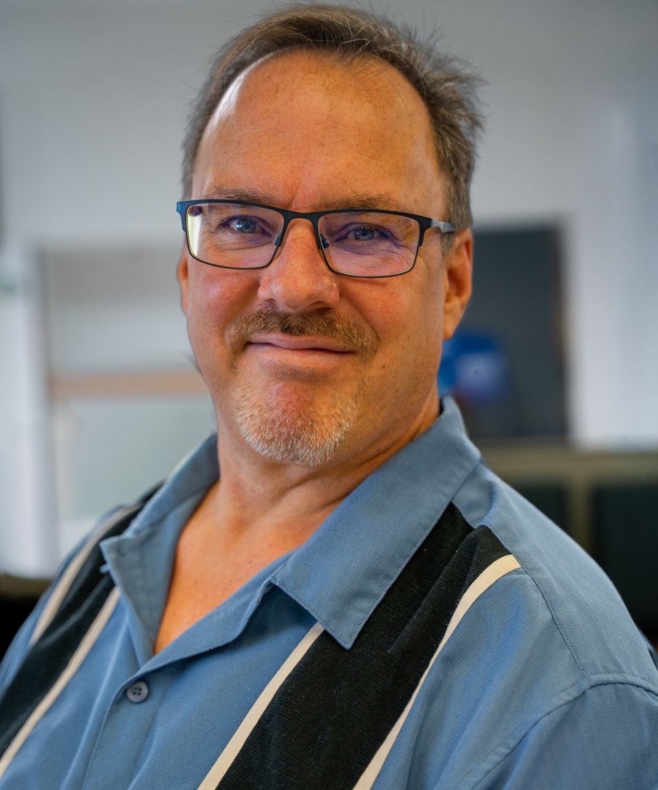A middle-aged man with glasses, facial hair, and a friendly expression, wearing a blue shirt with black suspenders, standing in an indoor setting.