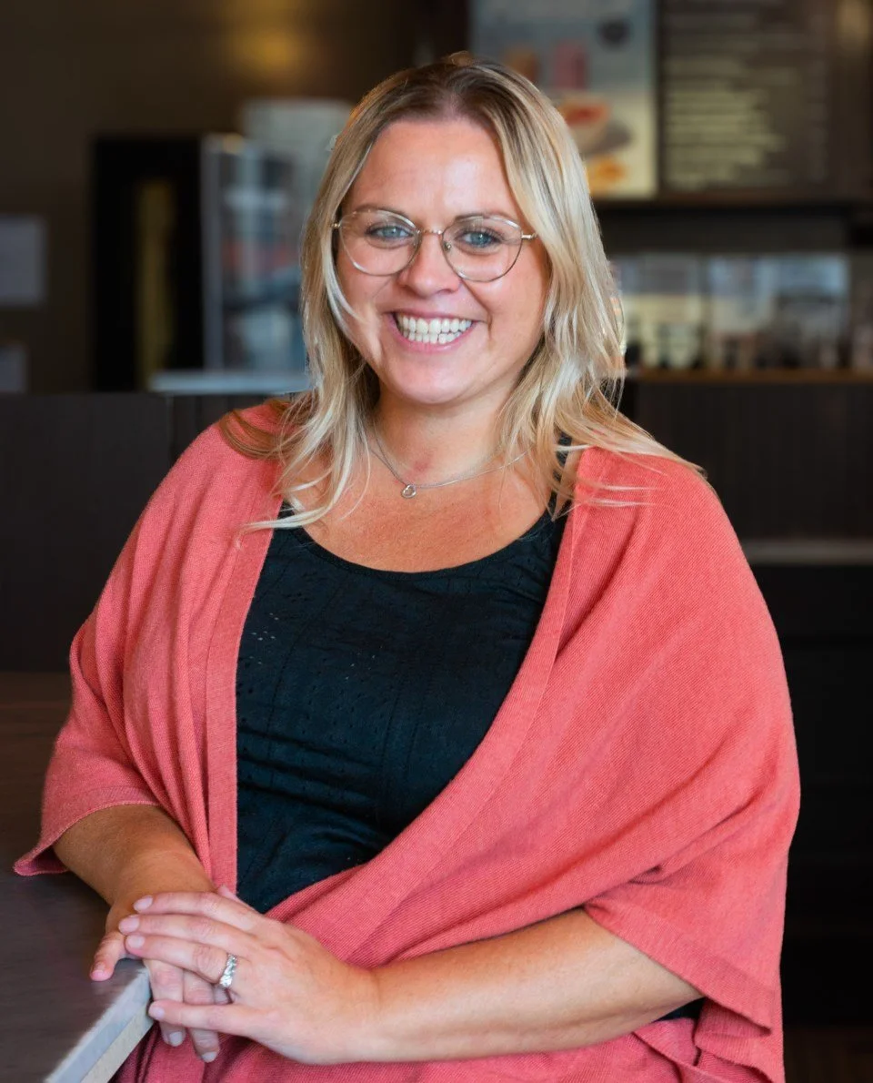 A woman with blonde hair and glasses smiling at a cafe, wearing a black top and a pink shawl.