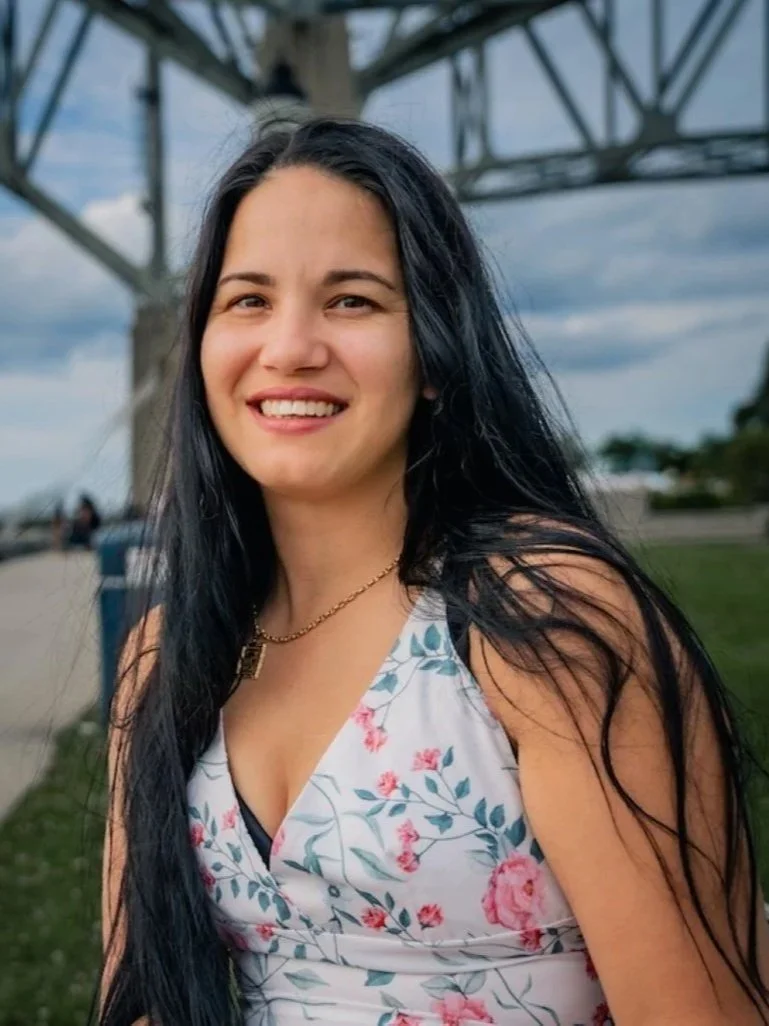 A young woman with long dark hair smiling outdoors near a metal structure and cloudy sky, wearing a floral sleeveless dress and gold necklace.