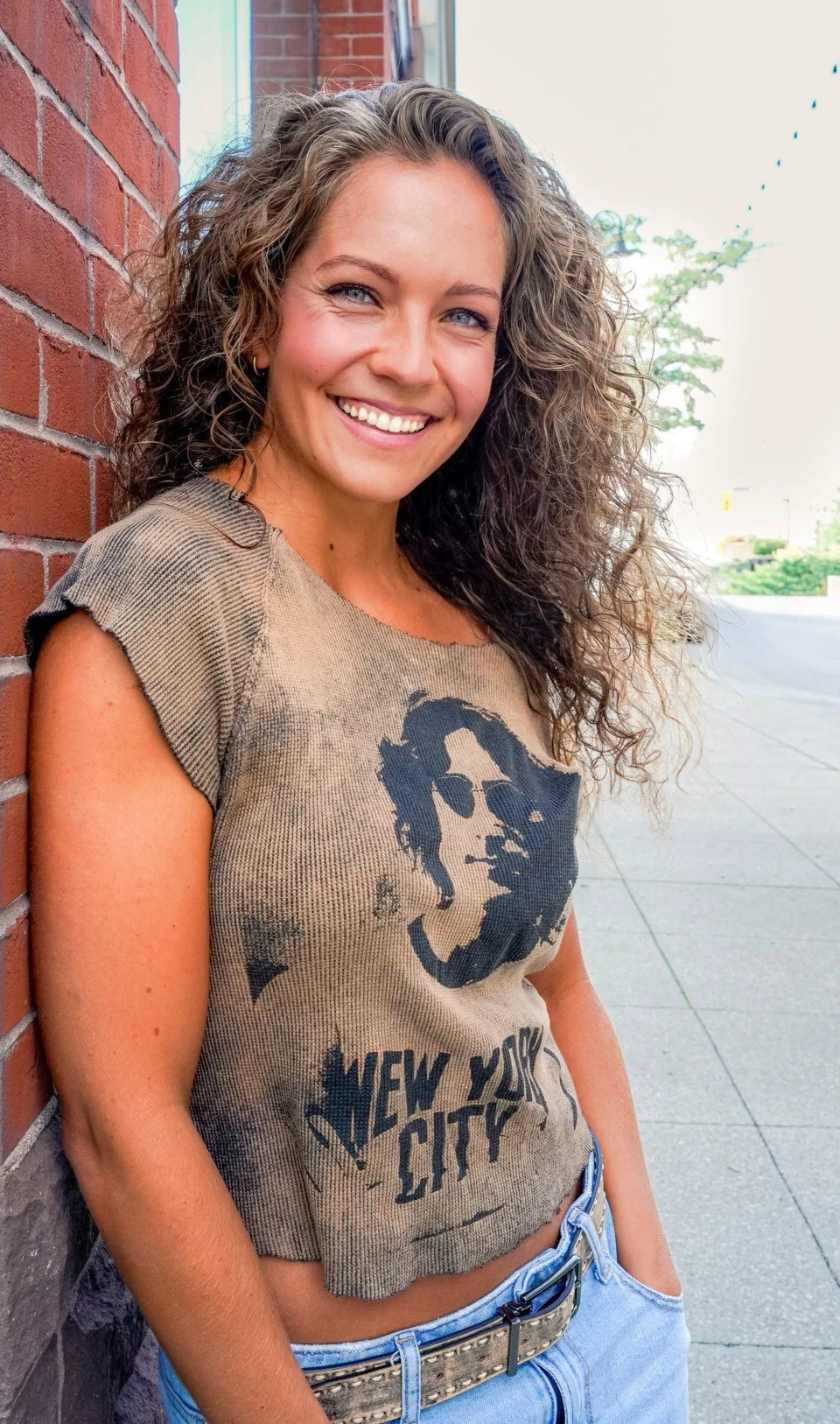 A smiling woman with curly hair leaning against a brick wall outdoors, wearing a beige T-shirt with a graphic of a man's face and the text 'New York City', and light blue jeans.