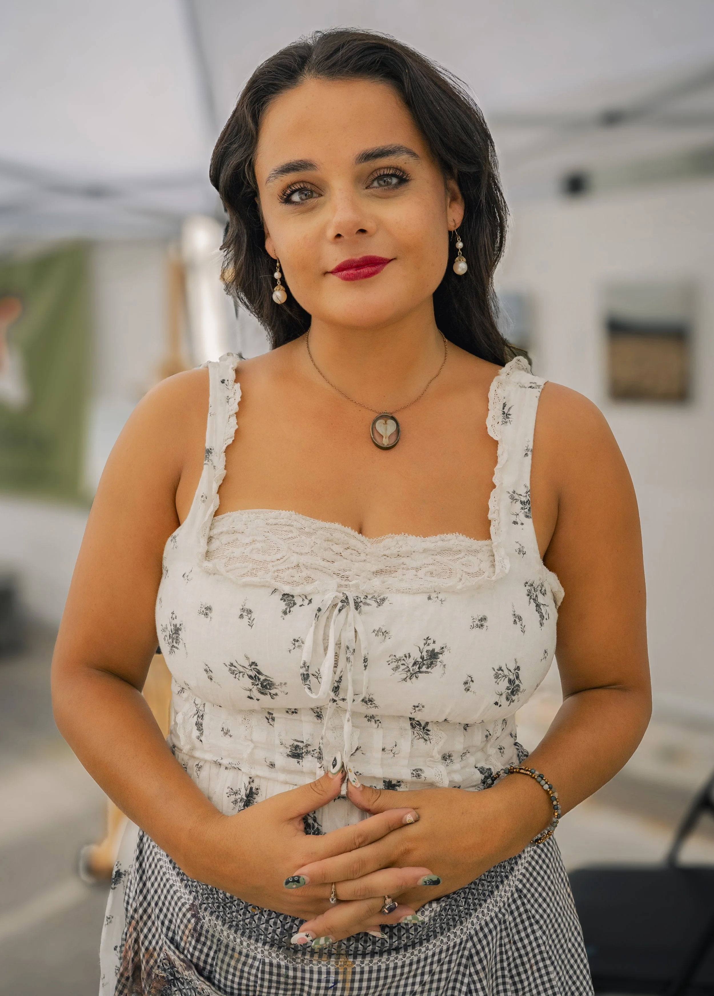 A woman with shoulder-length dark hair and light skin, wearing earrings and a necklace, standing indoors in front of a blurred background. She is dressed in a white sleeveless top with black floral prints and a checkered skirt, with her hands resting on her stomach.