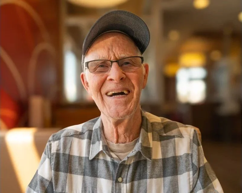 An elderly man with glasses and a baseball cap, wearing a plaid shirt, making a funny face in a cozy indoor setting.