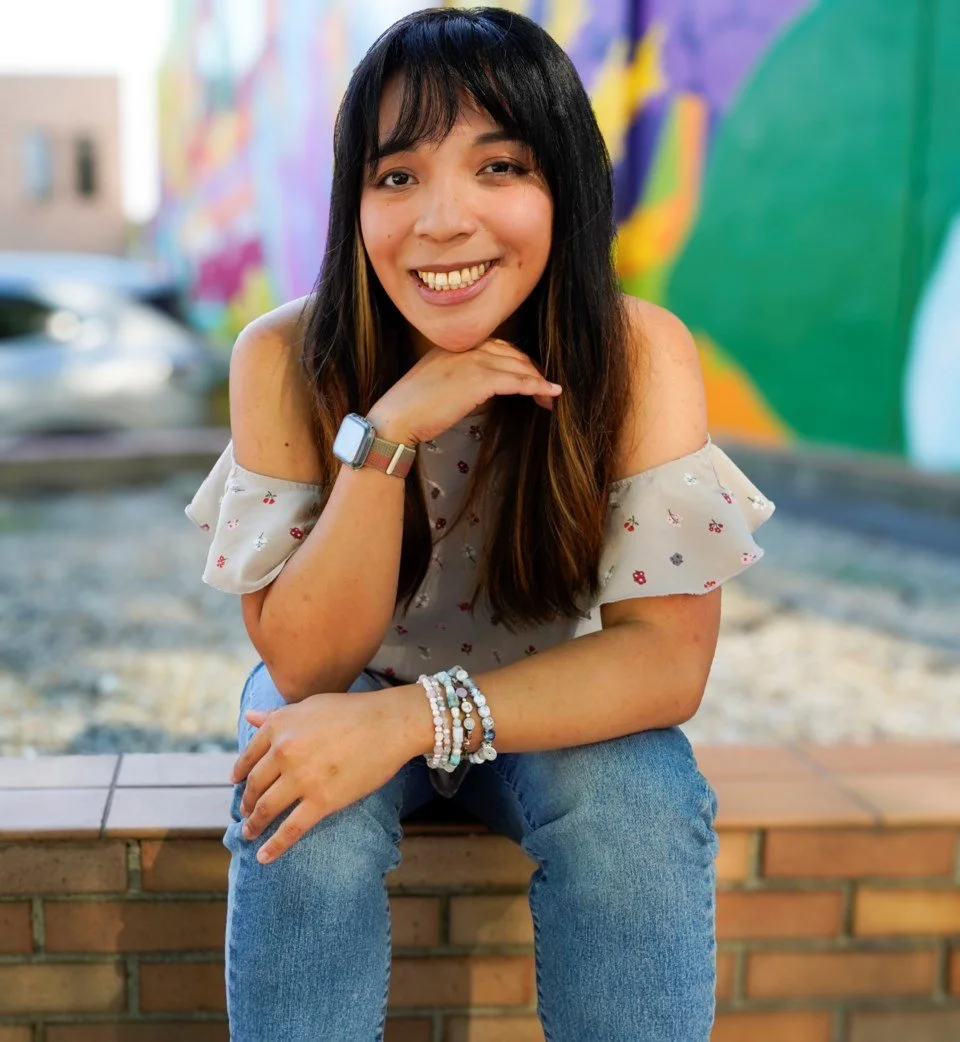 A smiling woman with long dark hair, wearing a light-colored off-the-shoulder top with small floral prints, blue jeans, and a smartwatch on her left wrist, sitting on a brick ledge in front of a colorful mural.