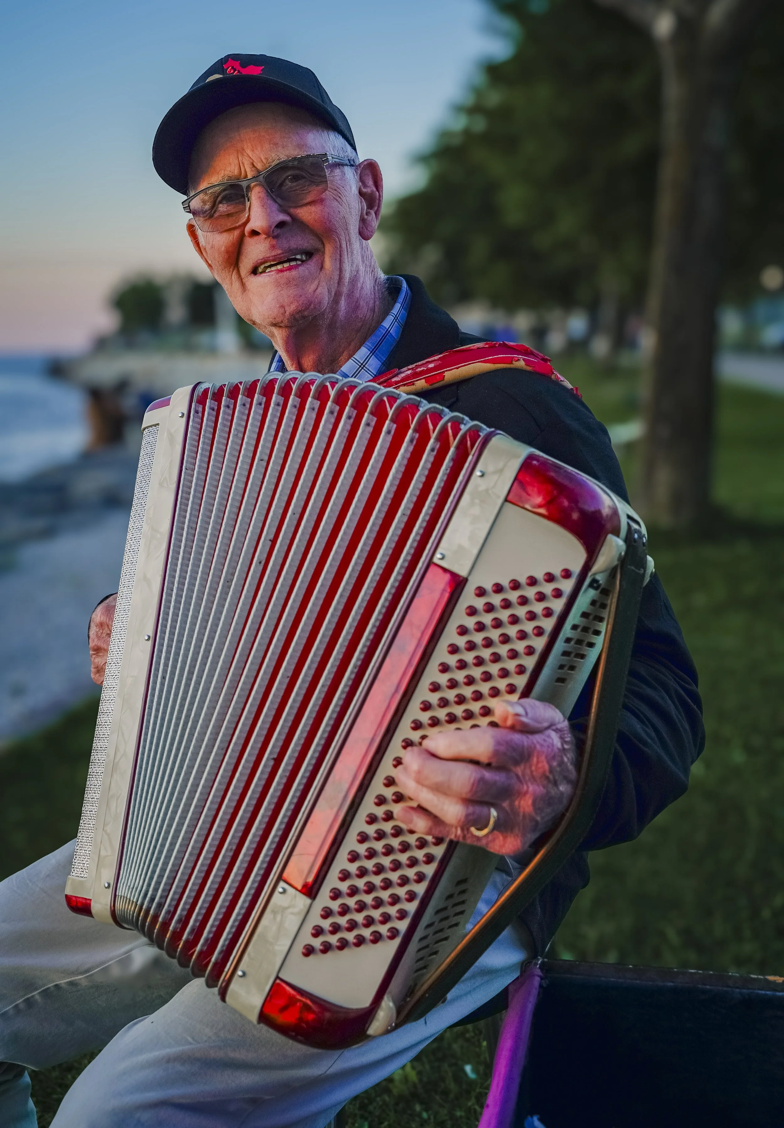 An elderly man wearing glasses and a black cap with a red logo, smiling while playing an accordion outdoors during sunset along a lakeside with trees in the background.