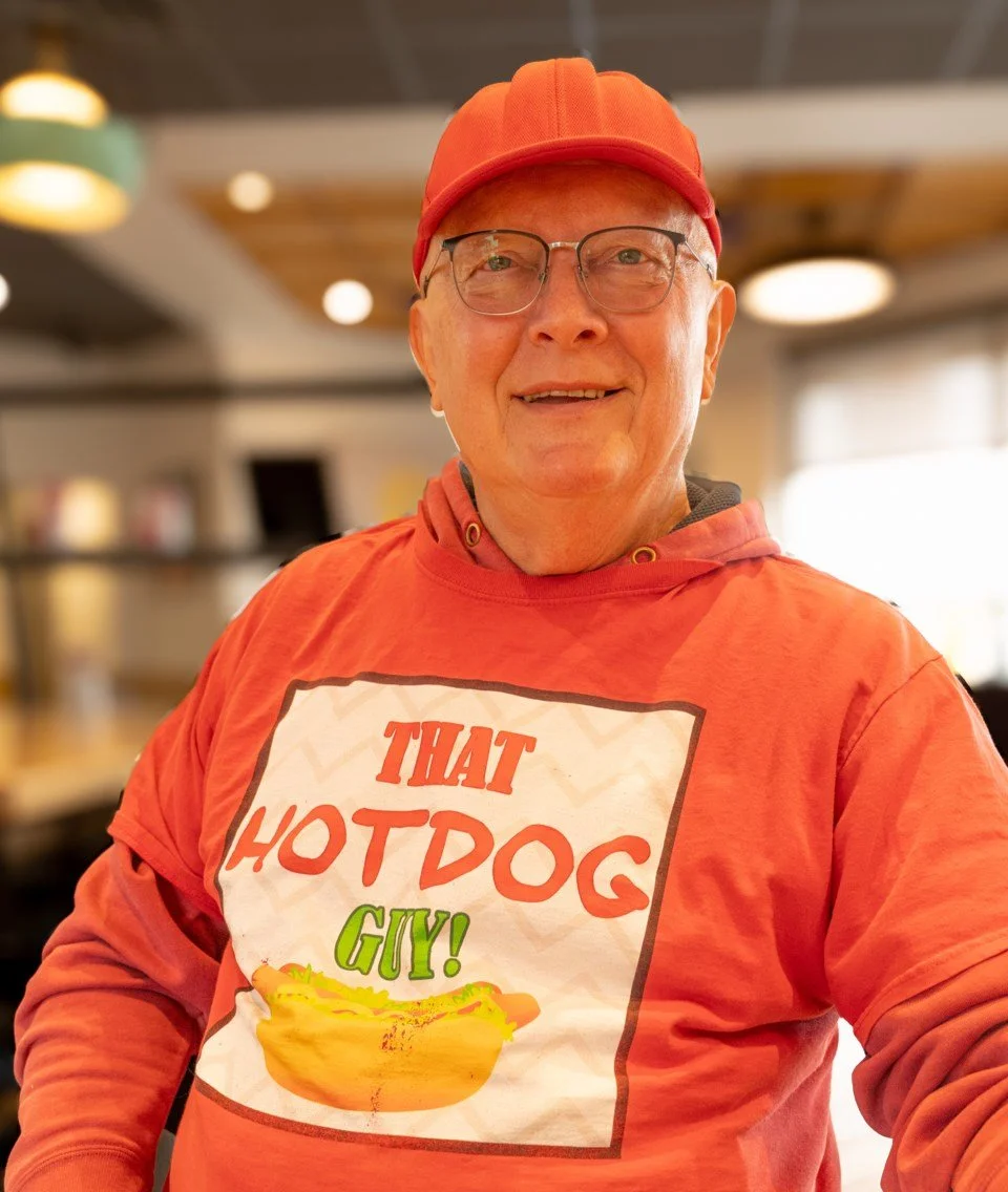 A man wearing glasses, a red cap, and a red hoodie with a graphic that reads 'That Hotdog Guy!' with a hotdog illustration, standing inside a restaurant.