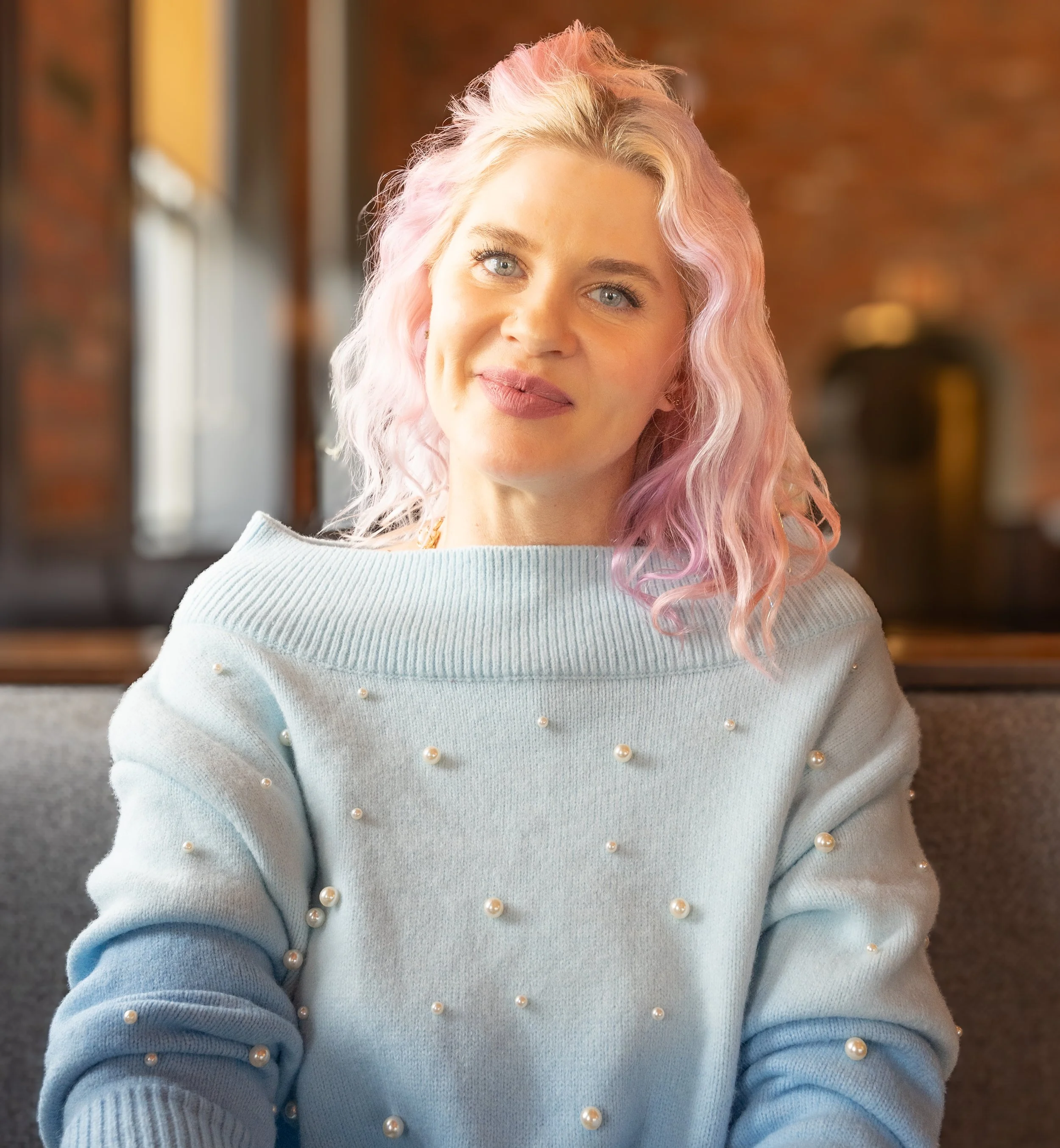 Close-up of a woman with pastel pink and blonde wavy hair wearing a light blue sweater with pearl embellishments.