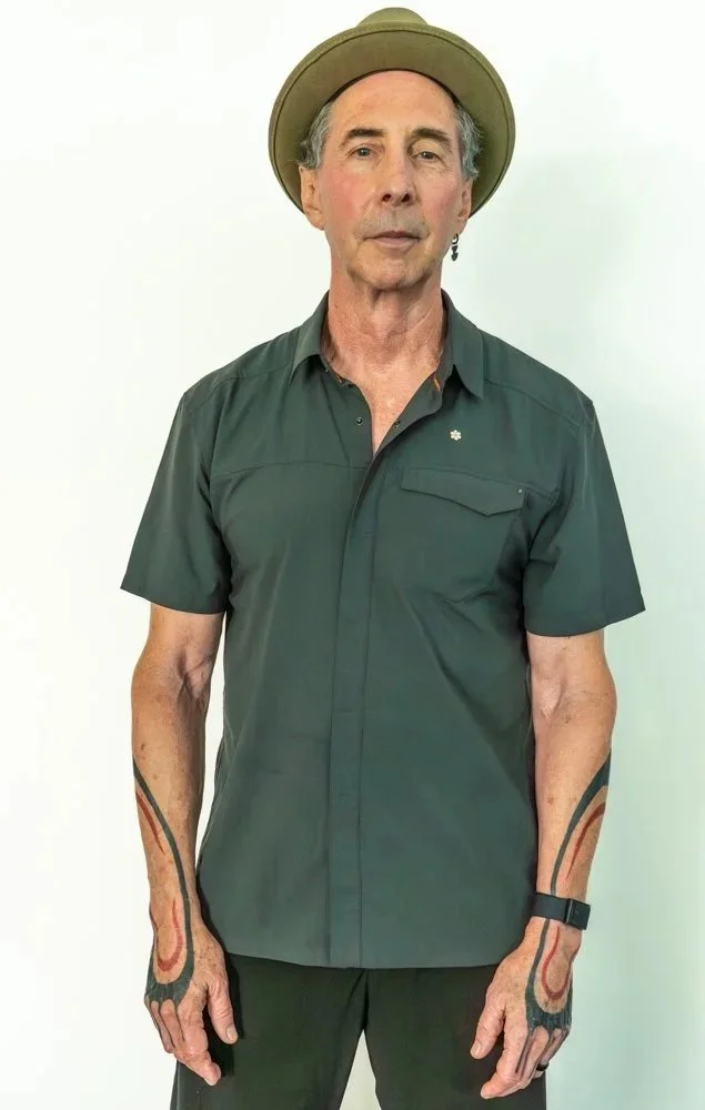 A middle-aged man wearing a wide-brimmed hat, dark green short-sleeve button-up shirt, and black pants. He has tattoos on both forearms, earrings, and a watch on his left wrist, standing against a plain white background.