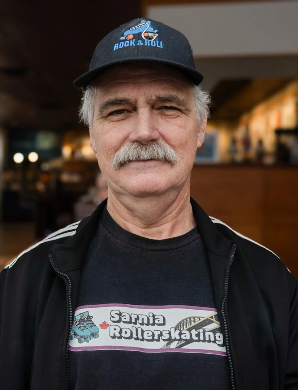 An elderly man with gray hair, mustache, and light skin, wearing a dark baseball cap with a graphic and the words 'Rock & Roll' on it, a black jacket with white stripes, and a black T-shirt with 'Sarnia Rollerskating' and an illustration of roller skates and a bridge. He is indoors in a restaurant or cafe.