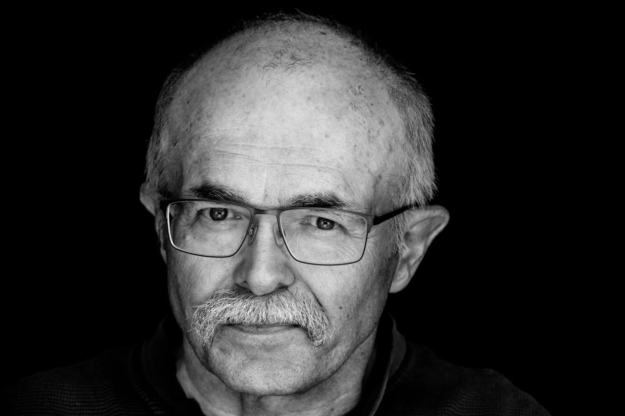 Close-up black-and-white photo of an elderly man with glasses, white hair, and a beard, facing forward with a neutral expression.