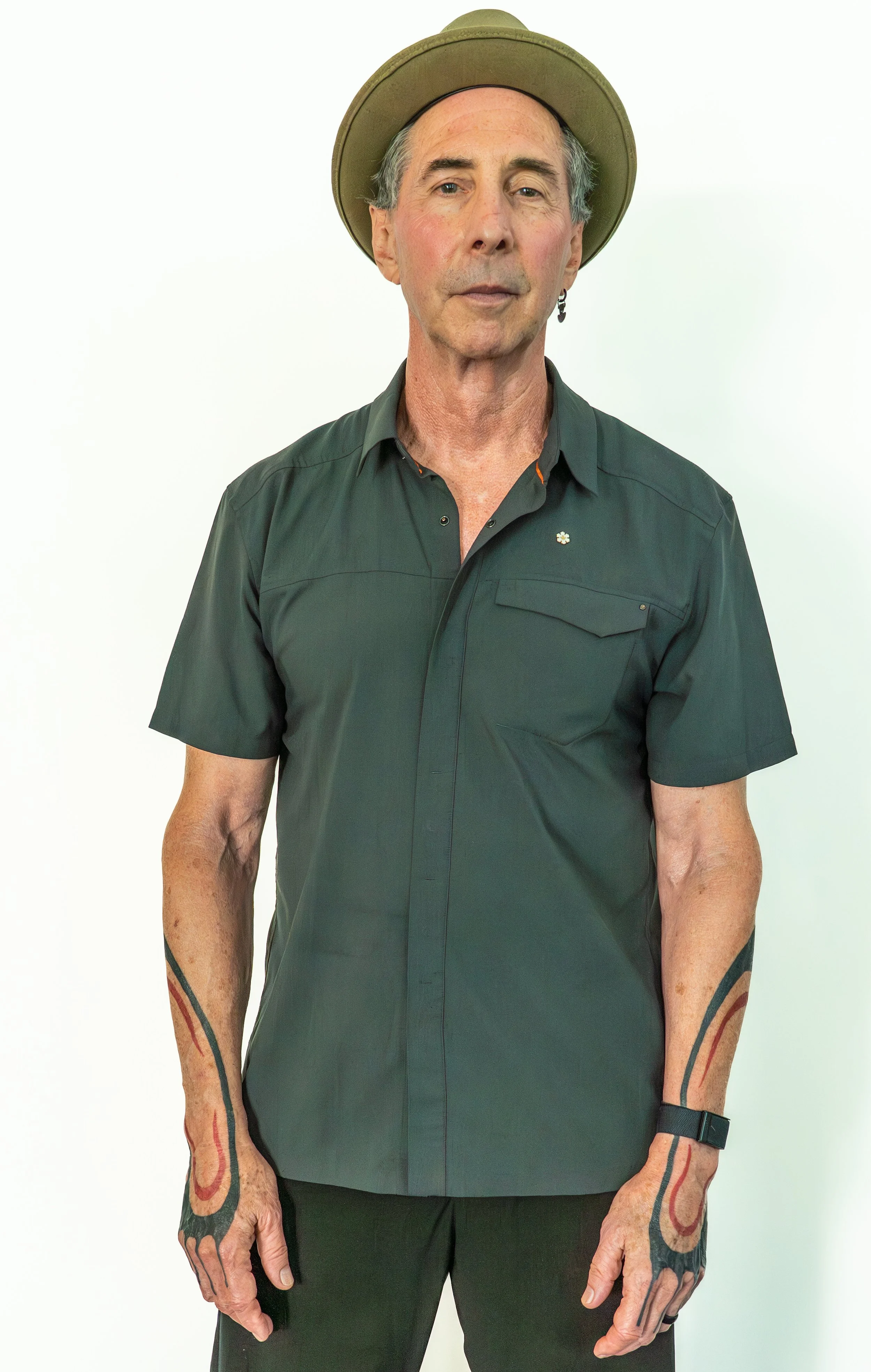 An older man with gray hair, wearing a brown wide-brimmed hat, a dark gray short-sleeved button-up shirt, and black pants, standing against a plain white background. He has tattoos on his arms, one of which is a red and black tattoo, and he is wearing an earring and a watch.