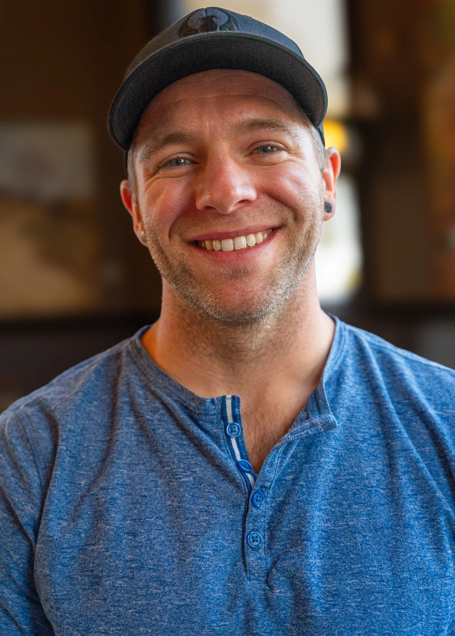 Close-up photo of a smiling man wearing a gray baseball cap with a bear logo, a blue button-up shirt, and earrings, with a blurred indoor background.