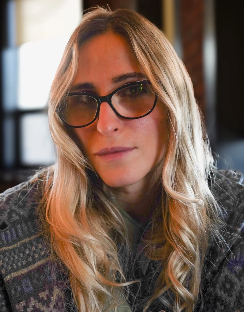 Close-up of a woman with long wavy blonde hair, wearing black sunglasses and a patterned sweater, indoors near a window.