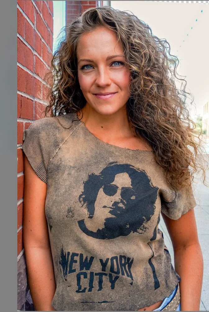 A young woman with curly hair, blue eyes, and light makeup, leaning against a brick wall, smiling at the camera, wearing a vintage-style t-shirt with a graphic of a person with sunglasses and the text 'New York City'.