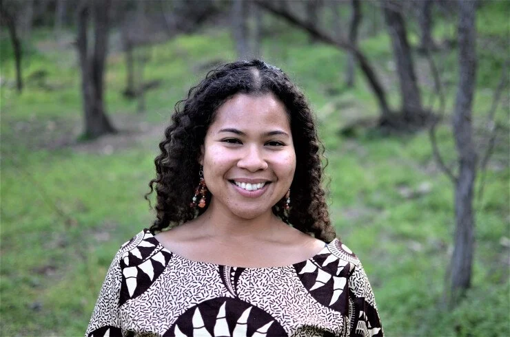 A woman with curly hair smiling outside in a green park with trees.