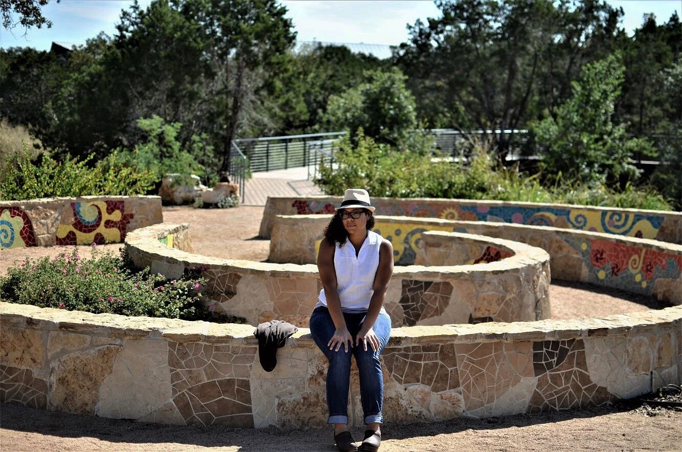 Woman sitting on a mosaic stone circular garden wall with colorful designs, wearing a white sleeveless blouse, jeans, a hat, and sunglasses, in a park or garden area with trees in the background.