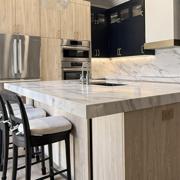 Modern kitchen with a marble island, light wood cabinets, dark upper cabinets, stainless steel appliances, and a marble backsplash.