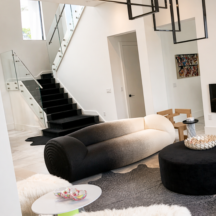 Modern living room with black and beige sofa, circular black ottoman, small white side table with bowls, abstract art on white walls, staircase with glass railing, and contemporary black lighting fixtures.
