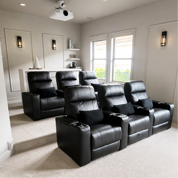 A home theater room with six black leather recliner seats arranged in two rows, black pillows on each seat, and a ceiling-mounted projector aimed at a blank wall.