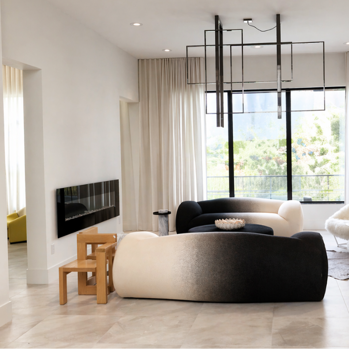 Modern living room with black and white curved sofas, a small wooden chair, and large windows with curtains overlooking trees, with a contemporary hanging light fixture.