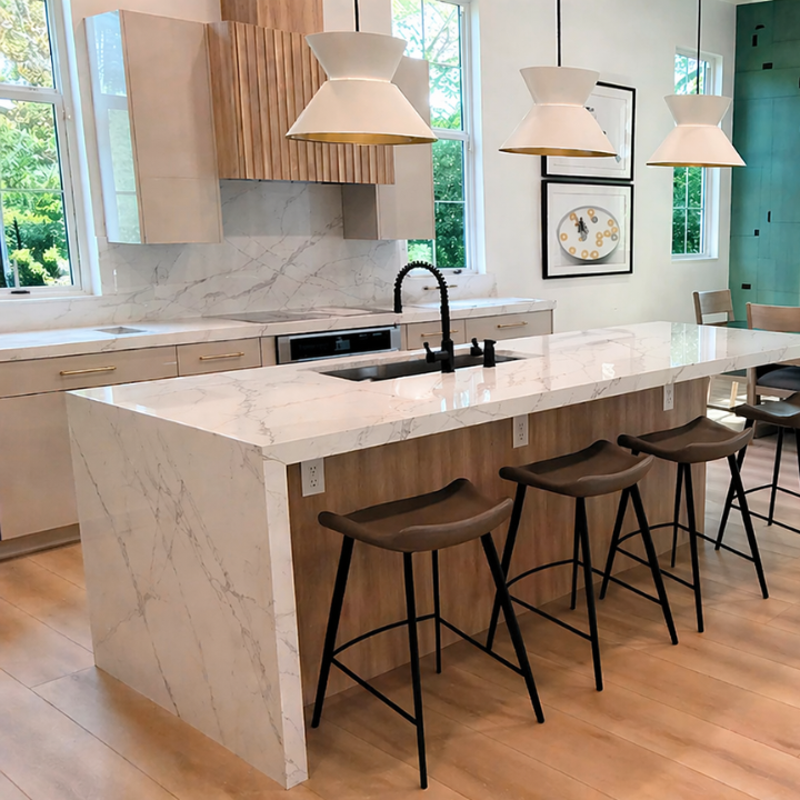 Modern kitchen with a white marble island, wooden bar stools, pendant lights, and a window view of greenery outside.