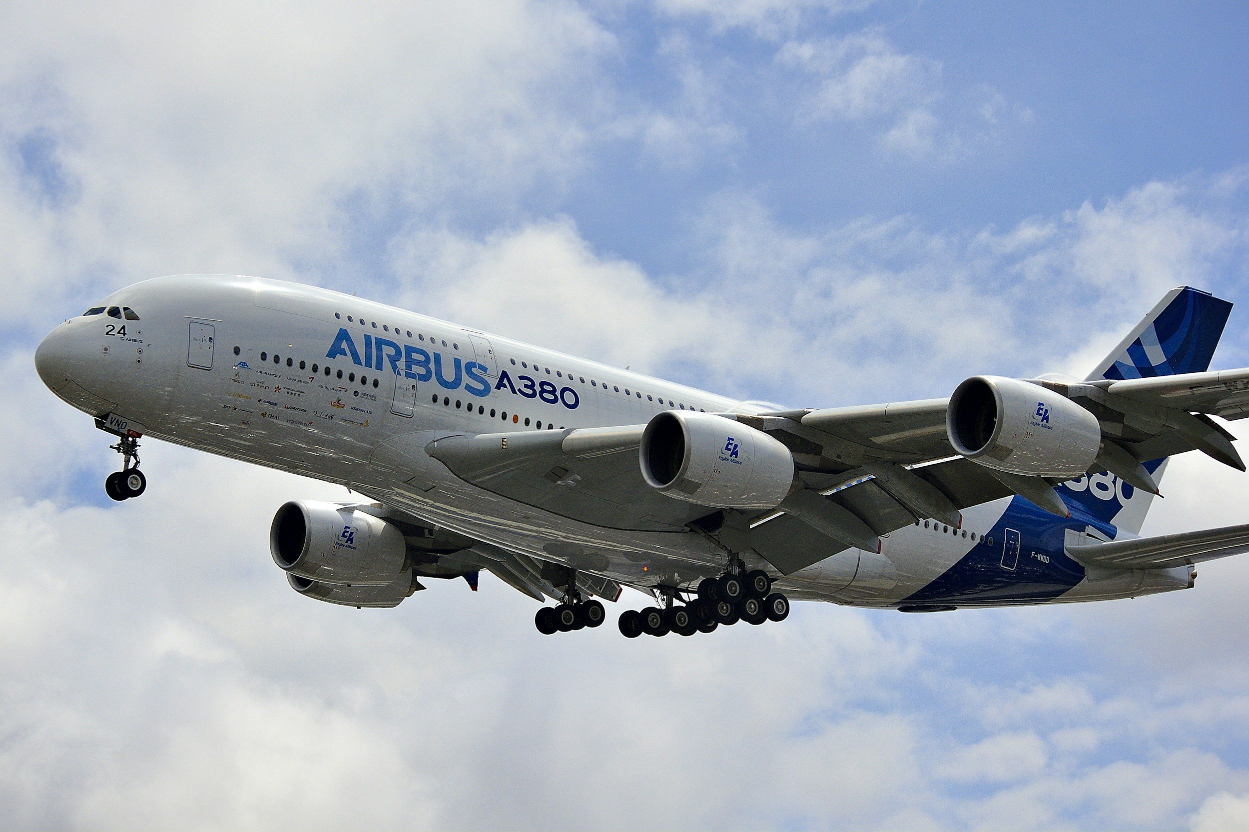 A white Airbus A380 airplane with blue branding flying in a partly cloudy sky.