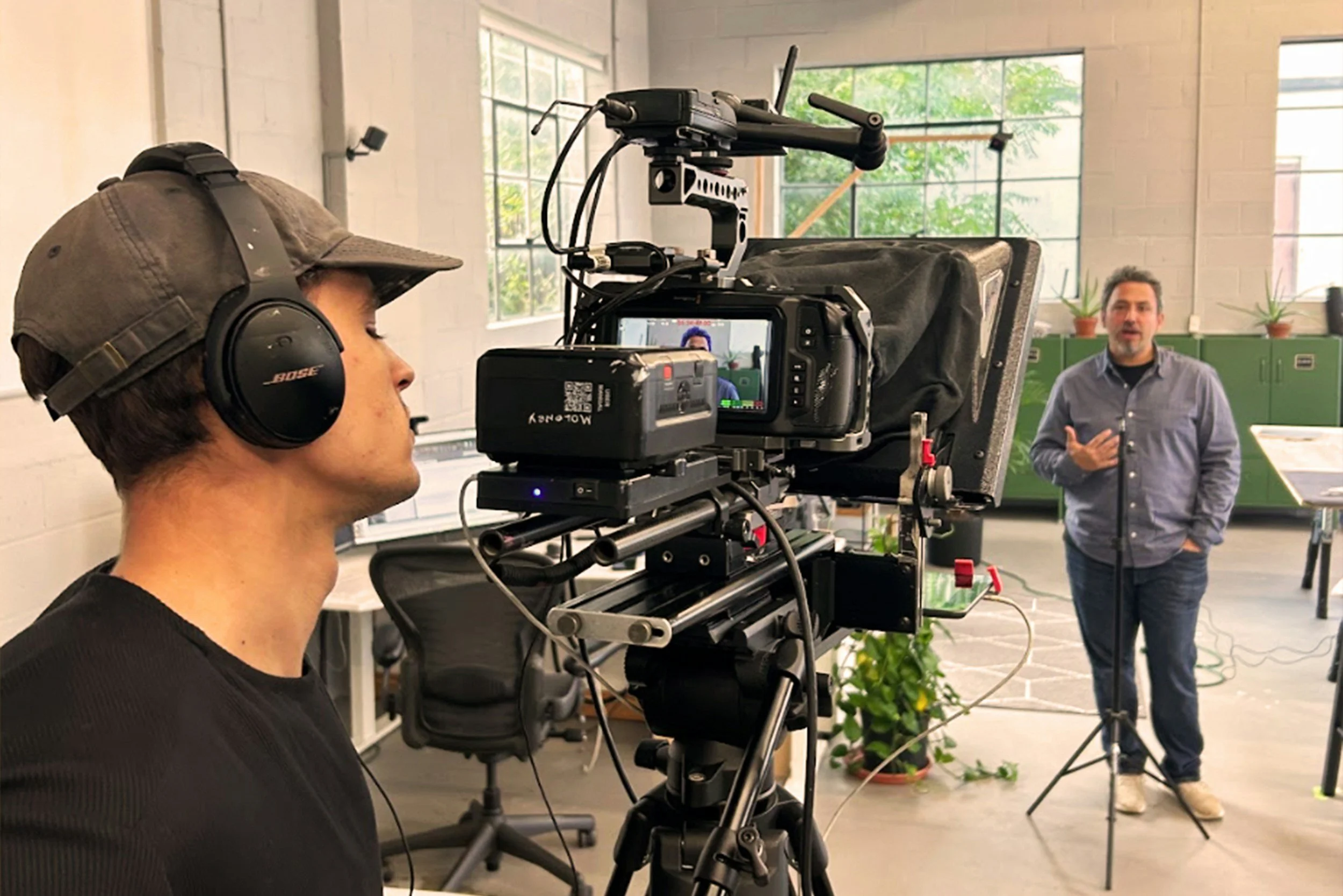 A person in a baseball cap and Bose headphones filming a man in a blue shirt giving a presentation in a studio with large windows and green cabinets.