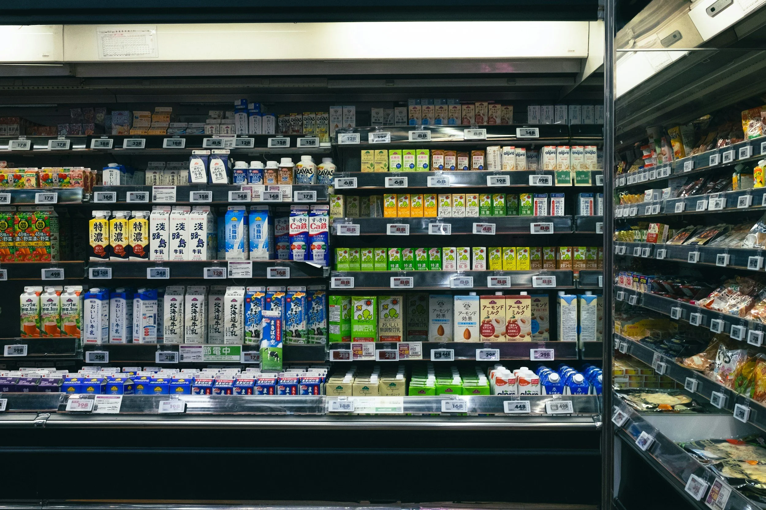 A grocery store refrigerated section with shelves stocked with various cartons of milk, juice, and dairy products, with price tags visible.