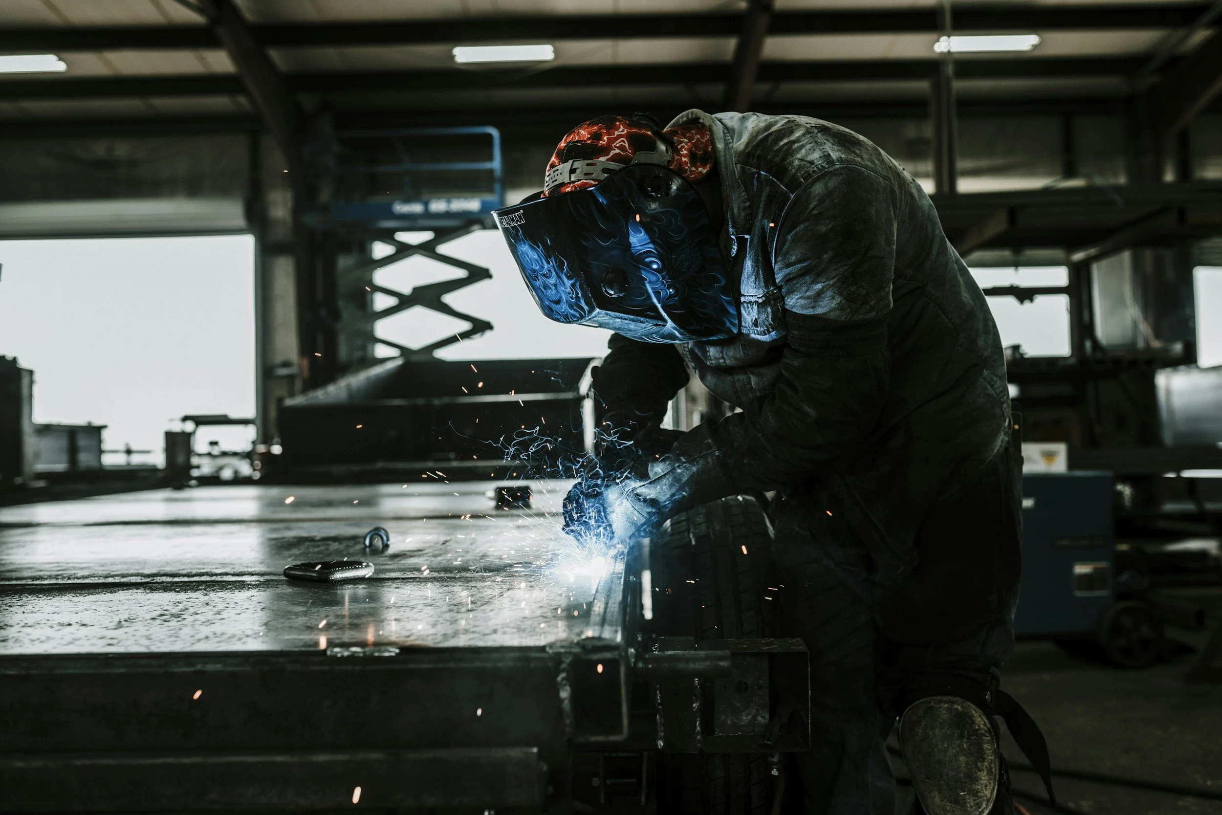A welder wearing a protective helmet and gloves welding metal on a workbench with sparks flying in an industrial workshop.