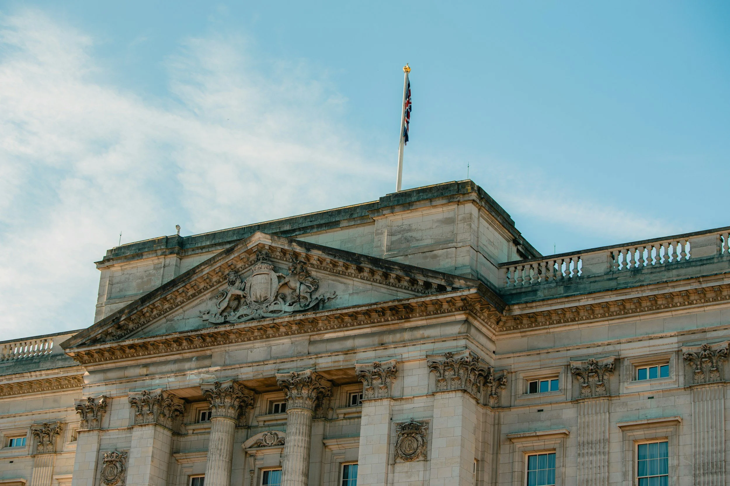 Close-up view of a historic government building with decorative columns and a crest, topped with a flagpole flying the Union Jack against a blue sky with clouds.