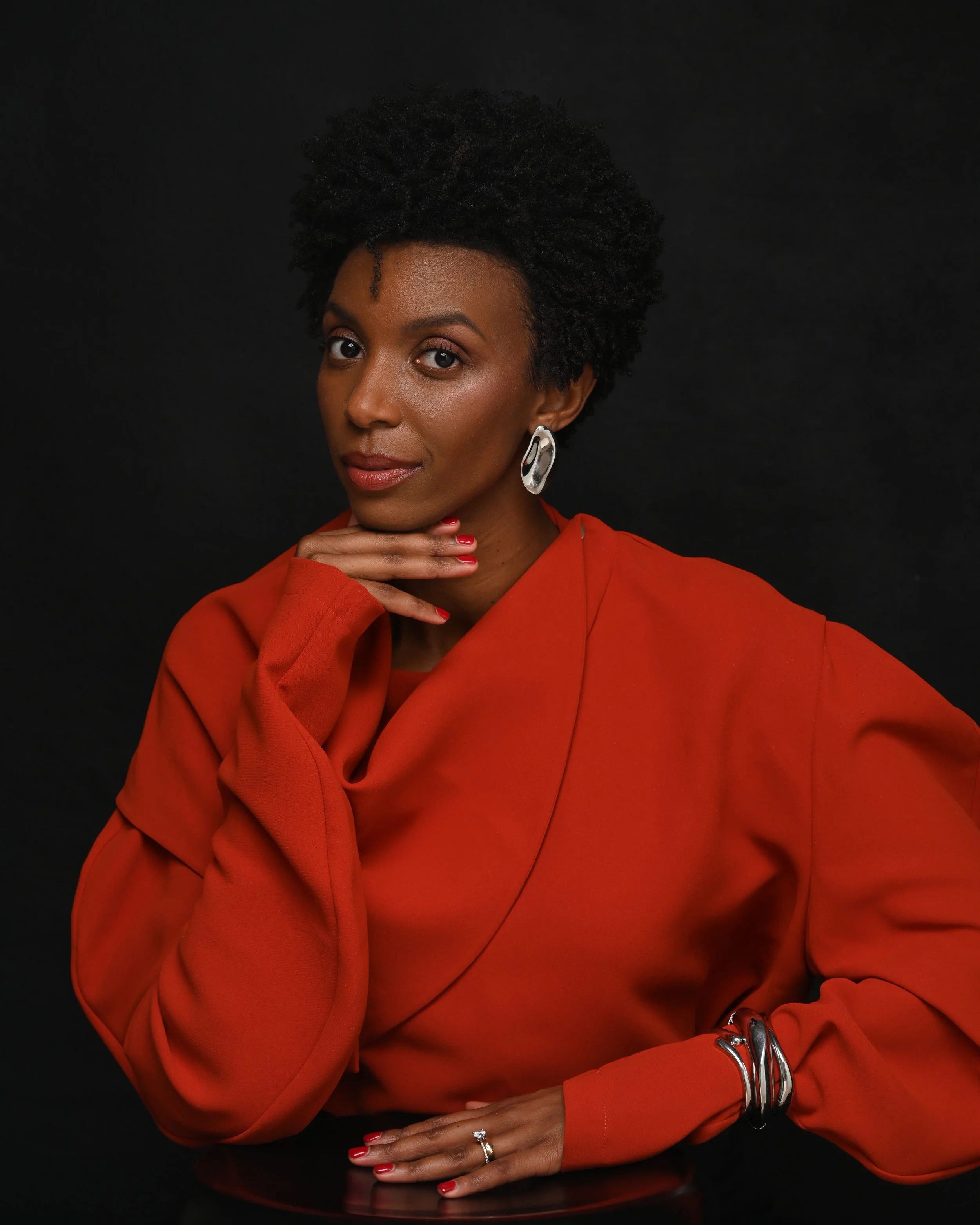 Portrait of a woman with short curly hair wearing a red blazer, silver jewelry, and red lipstick, posed with her hand under her chin against a black background.